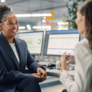 Two Female Colleagues Fondly Talk to Each Other, Laugh and Smile while Working on Computers in Diverse Modern Business Office. Experienced Manager and Young Employee Discuss a Fun Analytical Project.